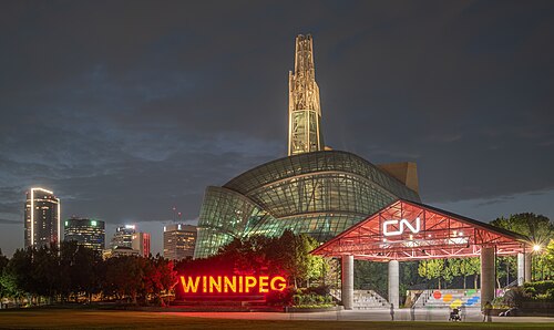 The Canadian Museum for Human Rights at night, viewed from The Forks in Winnipeg, Manitoba, Canada, with its illuminated glass atrium and spire rising above the downtown skyline. In the foreground, the illuminated "WINNIPEG" sign and the CN stage are visible.